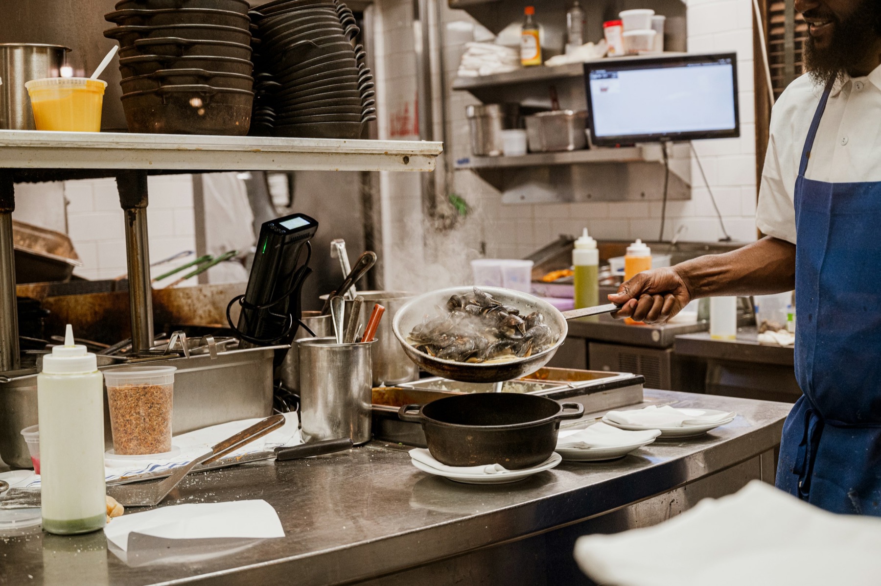 Een kok schept mosselen op in een restaurantkeuken, met het bestelscherm aan de muur