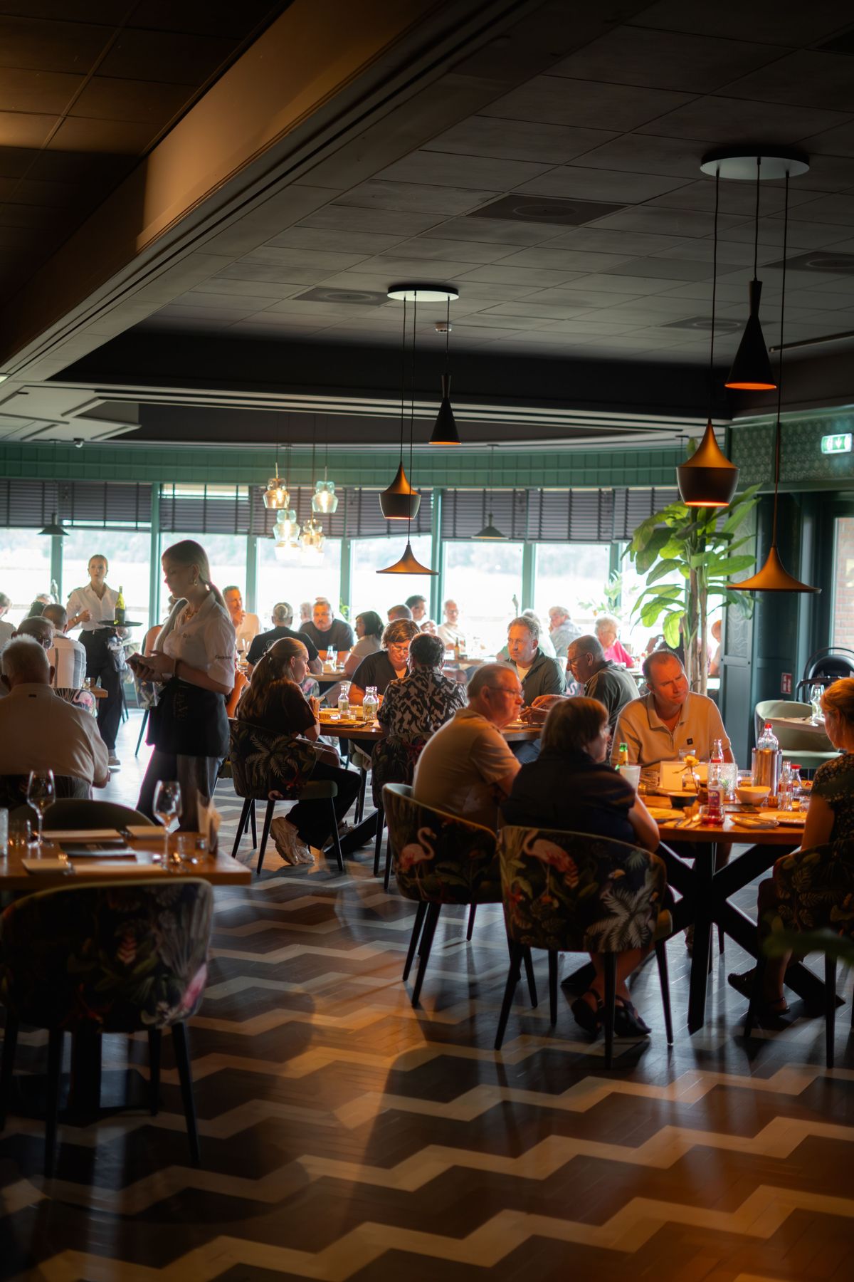 Volle restaurantzaal met grote ramen en gasten aan tafel